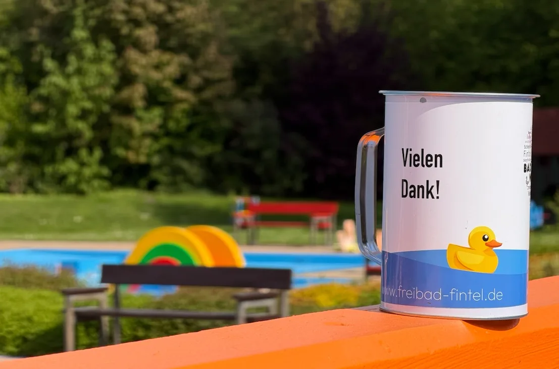 Spendendose 'Vielen Dank!' auf einer orangefarbenen Bank am Beckenrand des Freibads Fintel, im Hintergrund das Kleinkindbecken mit der bunten Rutsche